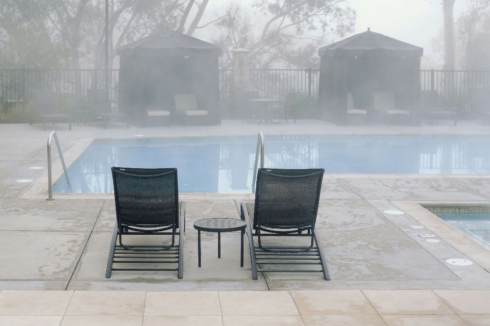 Two sling chaise lounges beside a foggy pool with cabanas in the background.